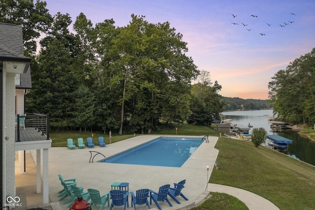 pool at dusk featuring a water view, a patio area, and a lawn