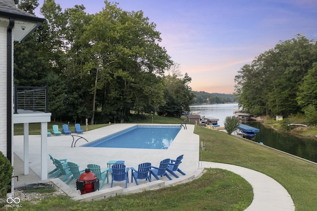 pool at dusk featuring a water view, a patio area, and a lawn