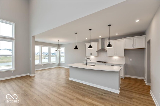 kitchen featuring a kitchen island with sink, white cabinets, wall chimney range hood, sink, and hanging light fixtures