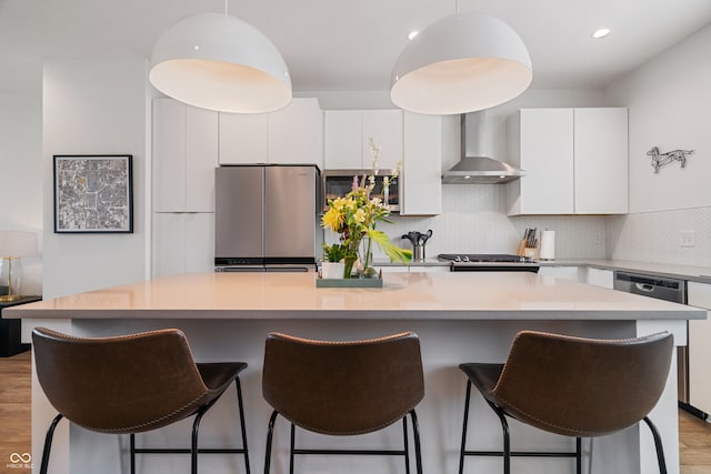 kitchen with appliances with stainless steel finishes, tasteful backsplash, a kitchen island, light wood-type flooring, and wall chimney exhaust hood