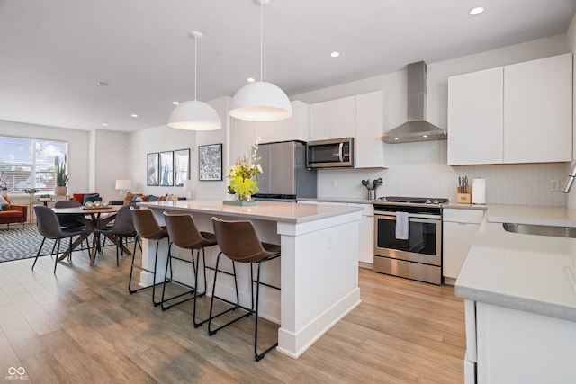 kitchen with white cabinetry, light wood-type flooring, wall chimney range hood, a center island, and appliances with stainless steel finishes