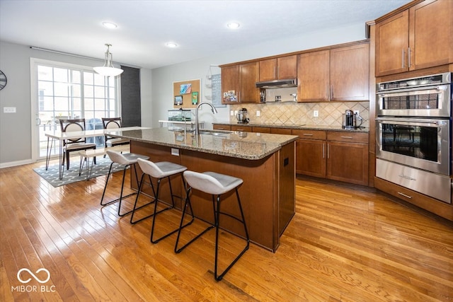 kitchen with a center island with sink, stainless steel double oven, hanging light fixtures, light hardwood / wood-style flooring, and sink