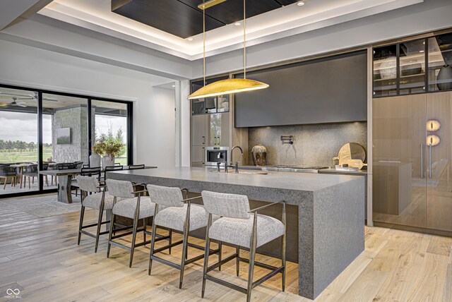 kitchen with a tray ceiling, tasteful backsplash, sink, hanging light fixtures, and light hardwood / wood-style flooring