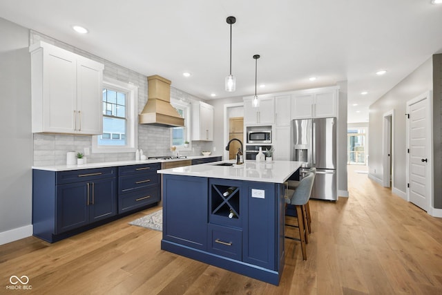 kitchen with premium range hood, blue cabinets, a center island with sink, stainless steel appliances, and white cabinets