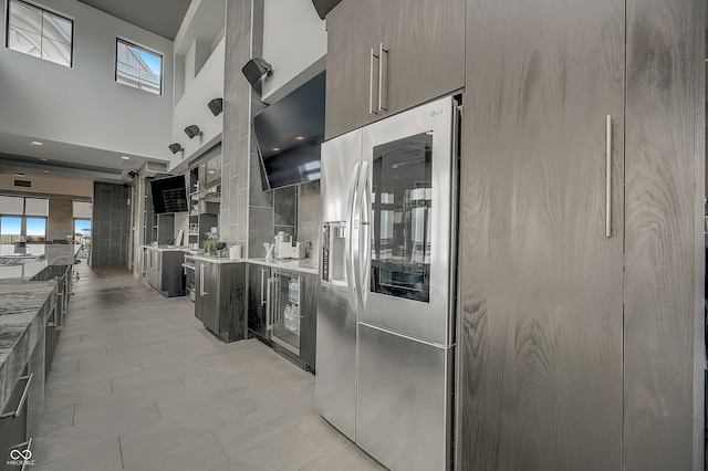 kitchen featuring light tile patterned floors, wine cooler, light stone countertops, a towering ceiling, and stainless steel refrigerator with ice dispenser