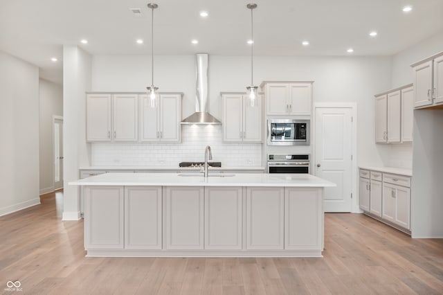 kitchen with an island with sink, wall chimney exhaust hood, hanging light fixtures, and appliances with stainless steel finishes