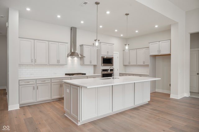 kitchen with wall chimney exhaust hood, an island with sink, decorative light fixtures, and stainless steel appliances