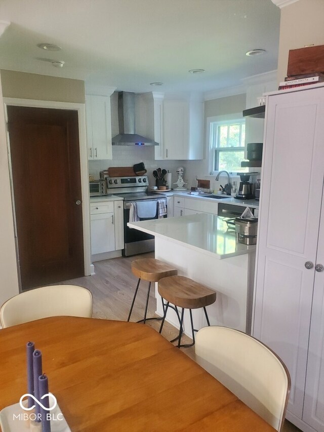 kitchen featuring white cabinets, stainless steel range with electric stovetop, a kitchen bar, light hardwood / wood-style floors, and wall chimney range hood