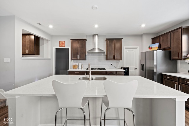 kitchen featuring wall chimney range hood, backsplash, dark brown cabinetry, a kitchen island with sink, and hardwood / wood-style flooring