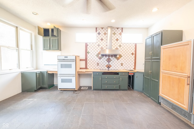 kitchen with white double oven, wall chimney exhaust hood, backsplash, a textured ceiling, and green cabinetry