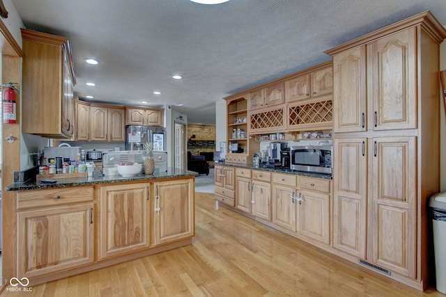 kitchen with light hardwood / wood-style flooring, stainless steel appliances, kitchen peninsula, light brown cabinets, and dark stone countertops