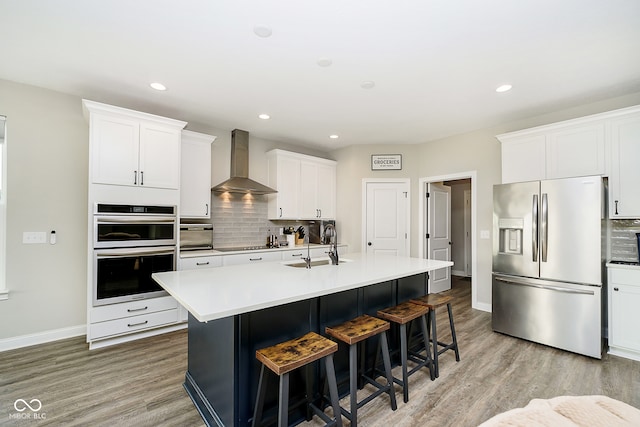 kitchen with wall chimney range hood, a kitchen island with sink, stainless steel appliances, white cabinets, and decorative backsplash