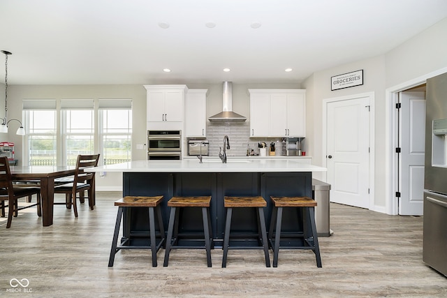 kitchen featuring white cabinets, decorative light fixtures, wall chimney exhaust hood, and a center island with sink