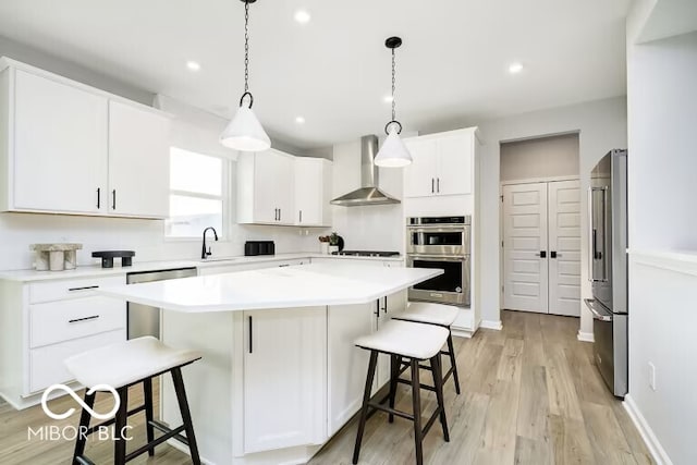 kitchen featuring wall chimney exhaust hood, appliances with stainless steel finishes, a kitchen bar, light wood-type flooring, and white cabinets