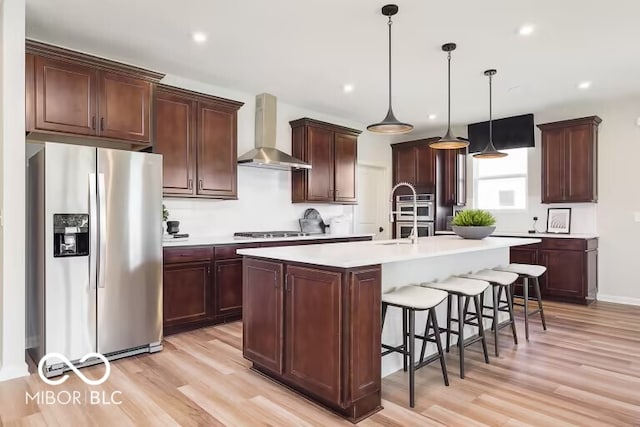 kitchen with hanging light fixtures, a center island with sink, wall chimney exhaust hood, stainless steel appliances, and light hardwood / wood-style floors