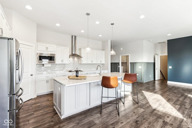 kitchen featuring wall chimney exhaust hood, hanging light fixtures, a center island with sink, white cabinetry, and appliances with stainless steel finishes