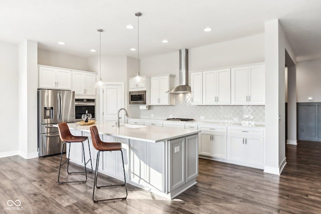 kitchen with wall chimney range hood, a kitchen island with sink, white cabinets, appliances with stainless steel finishes, and sink