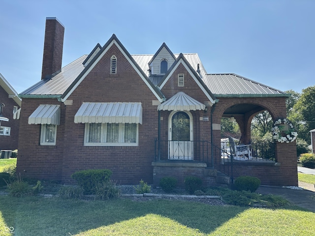 tudor-style house featuring brick siding, metal roof, a chimney, and a front lawn