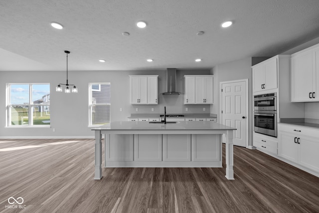 kitchen with dark wood-type flooring, stainless steel double oven, hanging light fixtures, and wall chimney exhaust hood