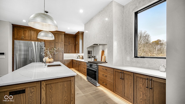 kitchen with brown cabinetry, a sink, an island with sink, high quality appliances, and light wood-type flooring