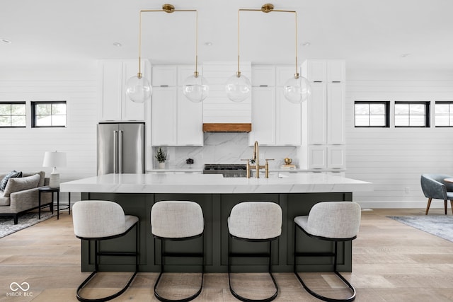 kitchen with light wood-type flooring, pendant lighting, a kitchen island with sink, stainless steel refrigerator, and wooden walls