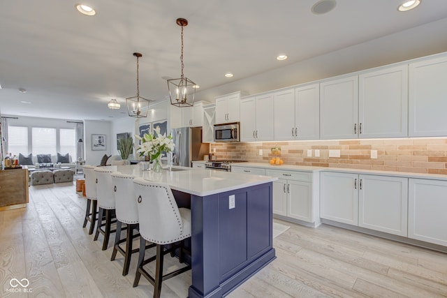 kitchen with light wood-type flooring, stainless steel appliances, a kitchen island with sink, white cabinetry, and hanging light fixtures