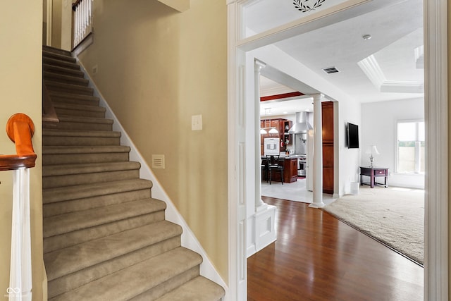 stairway featuring wood finished floors, visible vents, a tray ceiling, carpet, and decorative columns