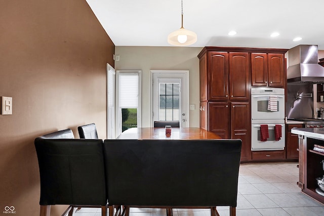 kitchen featuring light tile patterned floors, white double oven, a center island, decorative light fixtures, and wall chimney range hood