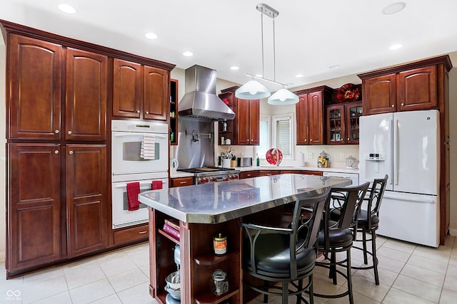 kitchen with a center island, white appliances, wall chimney exhaust hood, light tile patterned flooring, and a kitchen breakfast bar