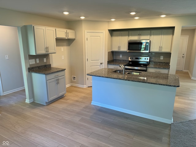 kitchen with appliances with stainless steel finishes, light hardwood / wood-style floors, dark stone countertops, and an island with sink