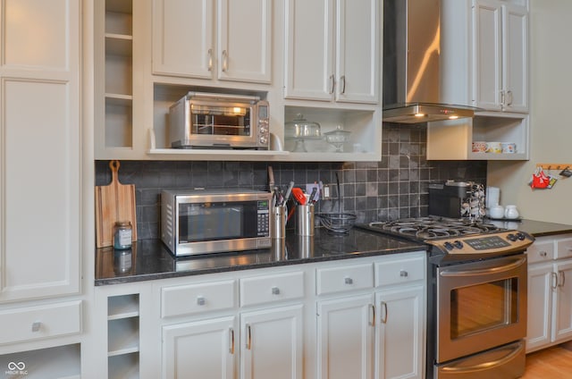 kitchen featuring dark stone counters, decorative backsplash, appliances with stainless steel finishes, white cabinets, and wall chimney range hood