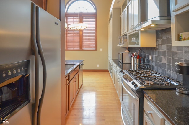 kitchen featuring dark stone counters, light hardwood / wood-style flooring, backsplash, wall chimney exhaust hood, and appliances with stainless steel finishes