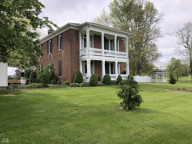 greek revival inspired property with a front lawn, a balcony, fence, covered porch, and brick siding