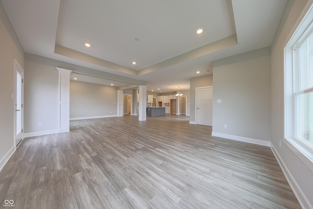 unfurnished living room featuring an inviting chandelier, a tray ceiling, light hardwood / wood-style floors, and decorative columns