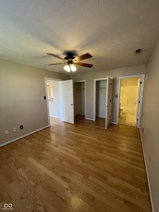 unfurnished bedroom with ensuite bath, a textured ceiling, light hardwood / wood-style floors, two closets, and ceiling fan