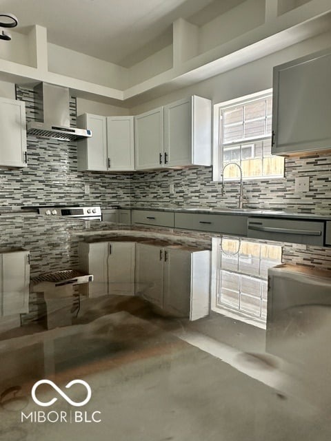 kitchen featuring concrete flooring, wall chimney range hood, white cabinetry, and backsplash