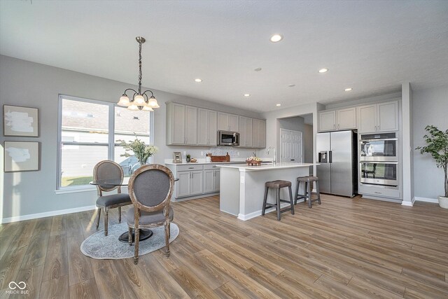 kitchen with gray cabinetry, a center island with sink, hanging light fixtures, stainless steel appliances, and light wood-type flooring