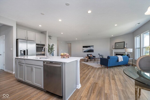 kitchen with gray cabinetry, a fireplace, light wood-type flooring, appliances with stainless steel finishes, and a center island with sink