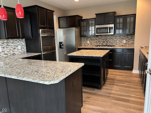 kitchen featuring light wood-style flooring, hanging light fixtures, appliances with stainless steel finishes, dark brown cabinets, and glass insert cabinets