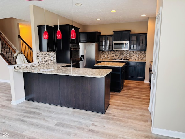 kitchen with decorative light fixtures, a peninsula, appliances with stainless steel finishes, light wood-type flooring, and glass insert cabinets