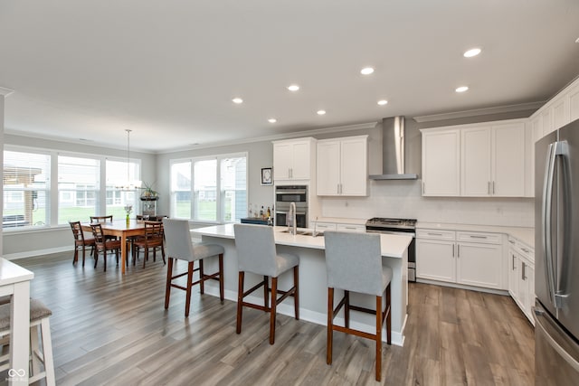 kitchen featuring pendant lighting, an island with sink, white cabinets, wall chimney range hood, and appliances with stainless steel finishes