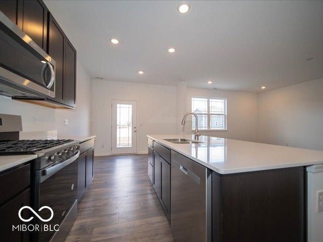 kitchen with a kitchen island with sink, a sink, light countertops, appliances with stainless steel finishes, and dark wood-style floors