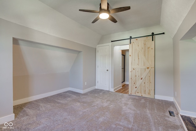 unfurnished bedroom featuring vaulted ceiling, a barn door, a textured ceiling, carpet flooring, and ceiling fan