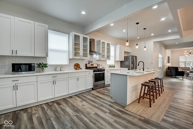 kitchen featuring a chandelier, an island with sink, white cabinets, wall chimney exhaust hood, and stainless steel appliances