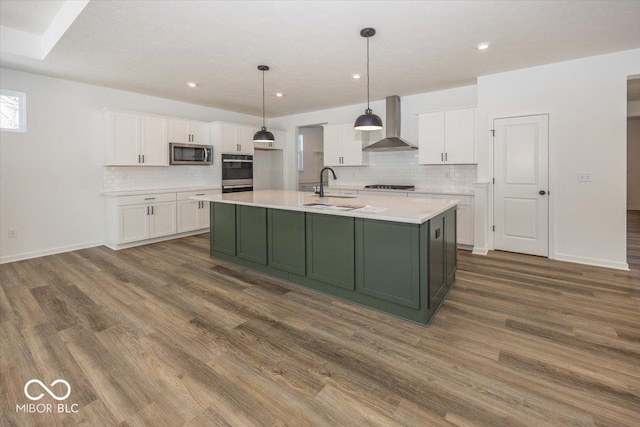 kitchen with white cabinets, a center island with sink, wall chimney exhaust hood, appliances with stainless steel finishes, and decorative light fixtures