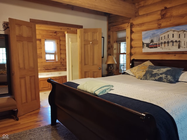 bedroom with light hardwood / wood-style floors, beamed ceiling, connected bathroom, and rustic walls