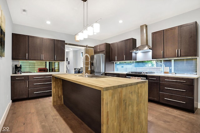 kitchen featuring a barn door, dark brown cabinetry, butcher block countertops, appliances with stainless steel finishes, and wall chimney exhaust hood