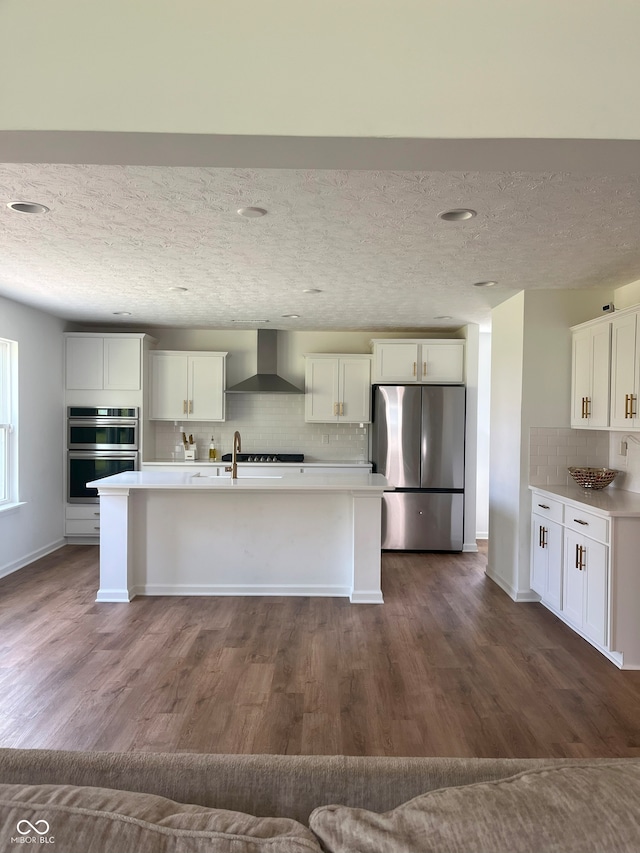 kitchen featuring appliances with stainless steel finishes, a center island with sink, dark hardwood / wood-style flooring, and wall chimney range hood