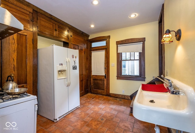kitchen featuring a sink, baseboards, white appliances, stone finish flooring, and wall chimney range hood