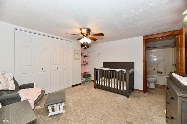 bedroom with a nursery area, ensuite bath, a textured ceiling, light colored carpet, and ceiling fan